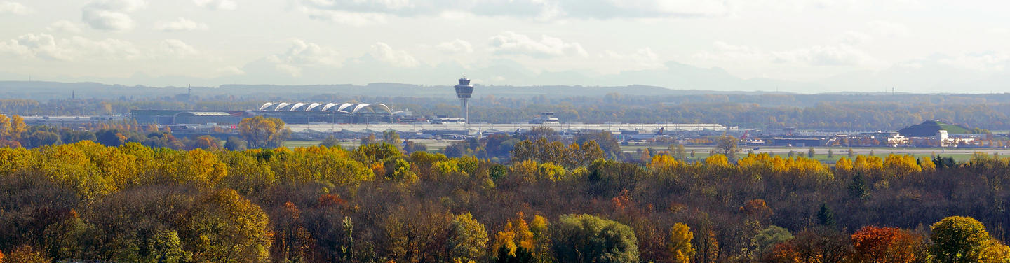 Panorama of the airport Panoramic view of Munich Airport behind autumn-colored trees, with terminals and the control tower set in open landscape and soft outlines of the Alps in the distance; calm and spacious atmosphere.