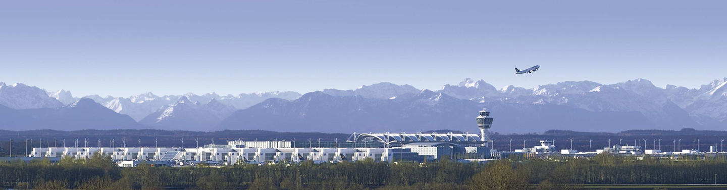 Munich Airport with Mountain Backdrop Panoramic image of Munich Airport with its tower and terminals in front of the Alps, with an airplane taking off in the background.