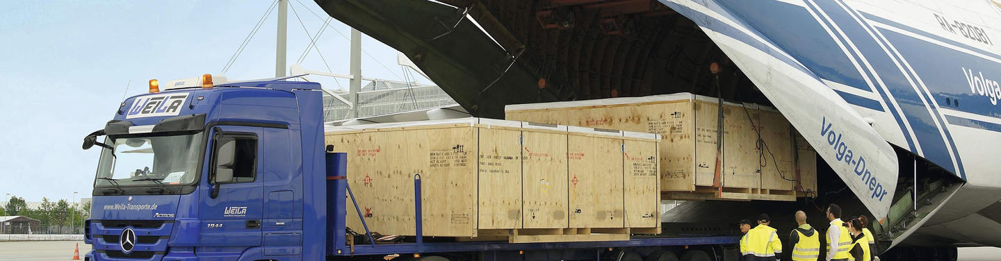 Tochtergesellschaft Cargogate Mitarbeitende in Warnwesten überwachen die Verladung großer Holzfrachtkisten aus einem Frachtflugzeug auf einen LKW am Cargogate des Flughafen München; konzentrierte, geschäftige Arbeitsatmosphäre.