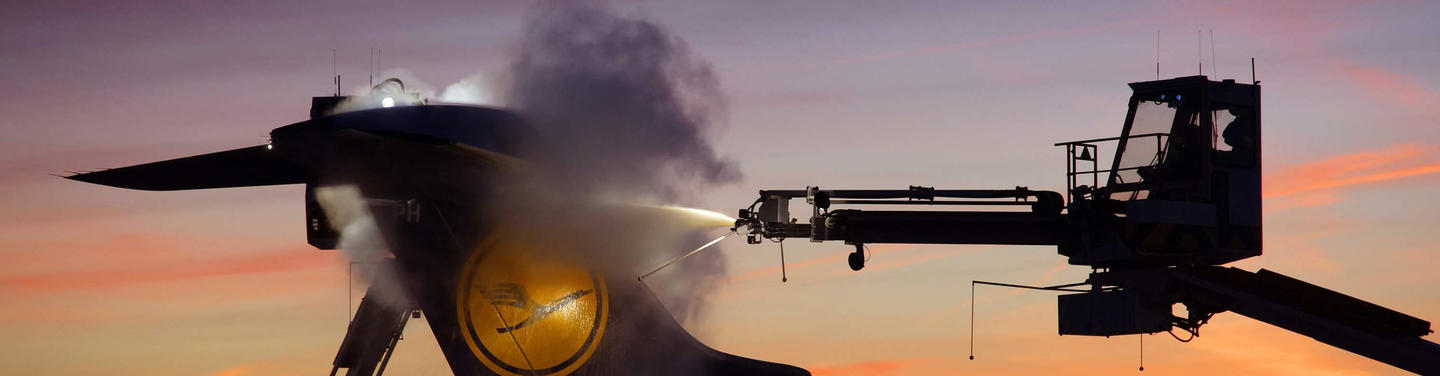 EFM Tochtergesellschaft Silhouette eines Enteisungsfahrzeugs sprüht Enteisungsflüssigkeit auf ein Flugzeug am Flughafen München; dampfende Wolken vor farbigem Abend- oder Morgenhimmel erzeugen eine ruhige, konzentrierte Einsatzstimmung.
