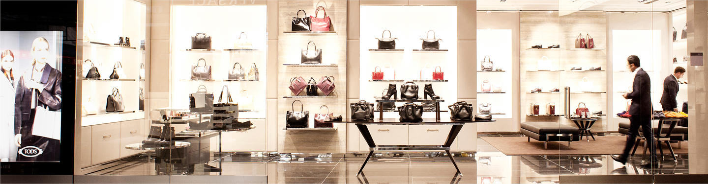 Tochtergesellschaft eurotrade Heller Eurotrade-Shop im Flughafen mit beleuchteten Regalen voller Handtaschen, Schuhe und Accessoires; ein Kunde läuft durch den modern eingerichteten Verkaufsraum mit glänzendem Boden.