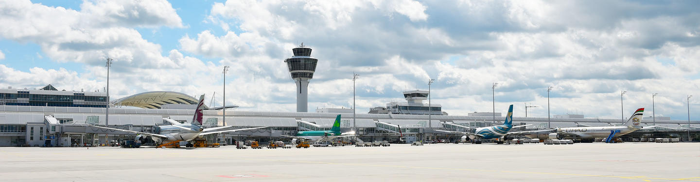 Flughafen München Weitläufige Ansicht des Vorfelds am Flughafen München mit mehreren parkenden Passagierflugzeugen, Terminalgebäuden und dem markanten Tower unter einem leicht bewölkten Himmel; die Stimmung wirkt ruhig, professionell, weltoffen und vermittelt die Dynamik eines internationalen Arbeitsplatzes.