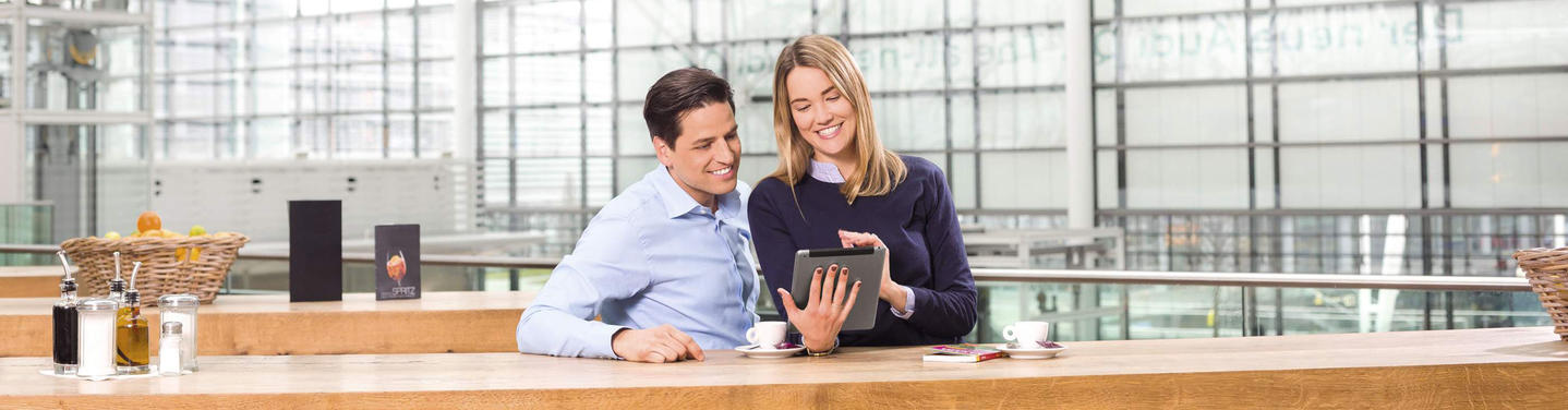 Service & Information at Munich Airport Two people sit at a counter in a modern airport setting and look at a tablet together, with cups and small items beside them; the scene represents service and information at Munich Airport and appears friendly and welcoming.