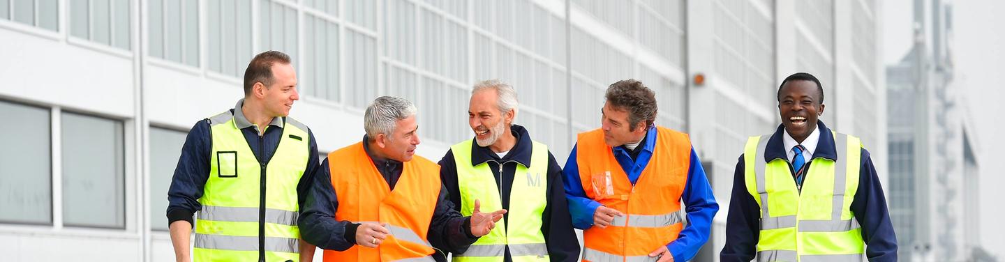 Teamgeist bei der AeroGround Fünf Männer in Warnwesten und Arbeitskleidung gehen lächelnd und im Gespräch nebeneinander an einem Flughafengebäude entlang. Die Stimmung wirkt freundlich, kollegial und teamorientiert.