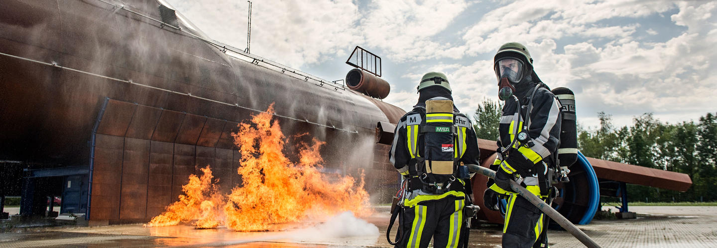 Seminarangebot Krisenmanagement der AirportAcademy - Flughafen München Mit Schutzanzügen ausgerüstete Flughafenfeuerwehrkräfte löschen einen großen Flammensimulator vor einer Übungsanlage. Die Szene verdeutlicht Einsatzbereitschaft, Training und höchste Sicherheitsstandards.