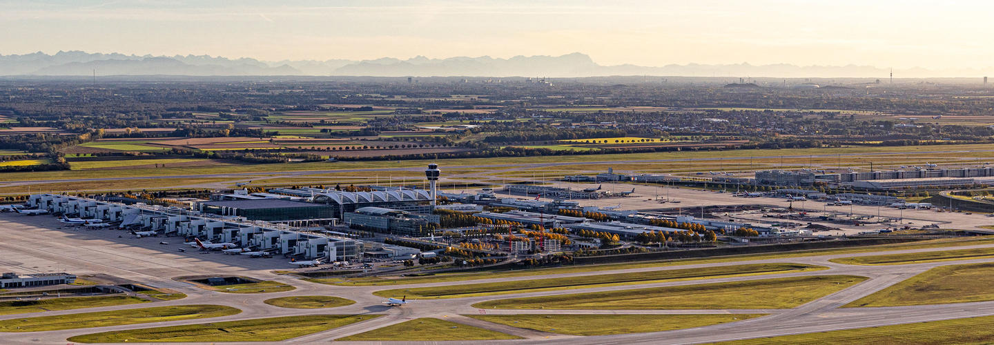 View at the alps Wide panorama of Munich Airport in the warm morning light, with terminals, runways, and the control tower in the background. The scene conveys the structure, tranquility, and size of this international hub.