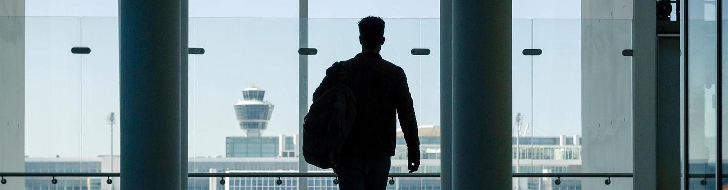 Kampagne "Where is Wayne" Silhouette einer Person mit Tasche, die in einer hellen Flughafenhalle vor großen Fenstern steht und auf das Vorfeld mit dem markanten Tower des Flughafens München blickt.