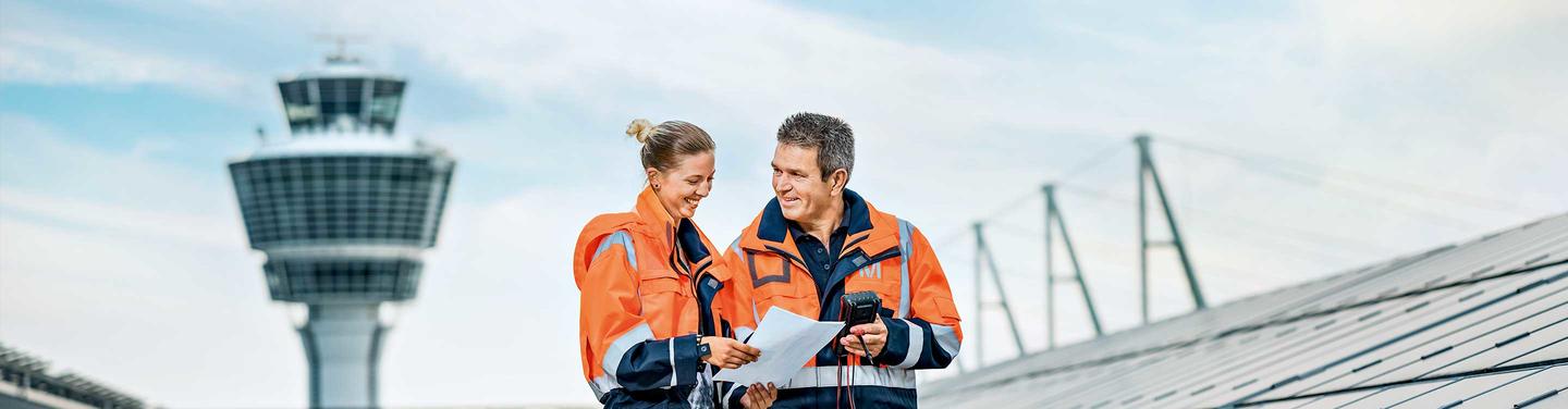 Duales Studium Bachelor of Engineering Elektro- und Informationstechnik Zwei Personen in orangefarbener Sicherheitskleidung stehen auf dem Flughafengelände vor dem Tower und besprechen technische Unterlagen; die Szene wirkt wie eine praxisnahe Arbeitssituation im technischen Bereich.