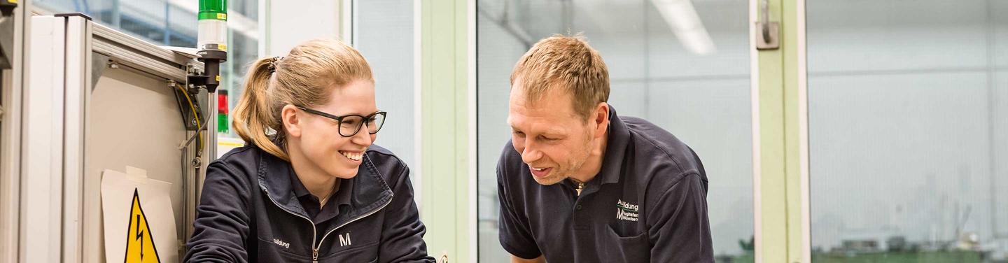 Ausbildung Mechatronik am Flughafen München Zwei Personen in Arbeitskleidung des Flughafens München arbeiten lächelnd und konzentriert an einer technischen Anlage; freundliche und kooperative Lernatmosphäre.