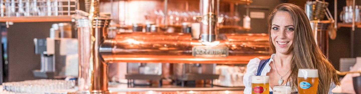 Airbräu im Flughafen München Mitarbeiterin in Tracht steht vor kupfernen Brauanlagen im Airbräu und hält mehrere frisch gezapfte Biergläser.