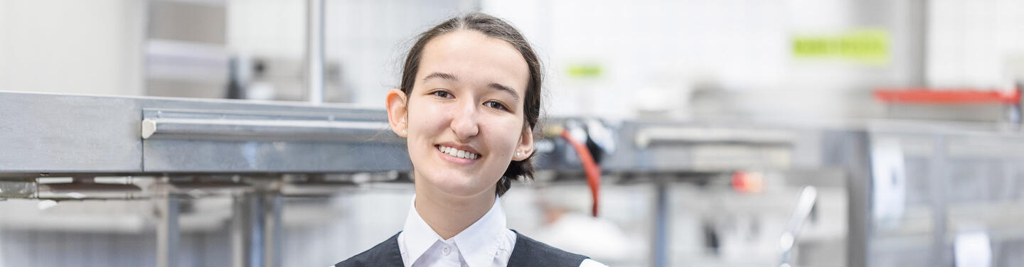 Allresto Azubi im Service Junger Mensch in Servicekleidung steht in einer Großküche und lächelt freundlich in die Kamera.