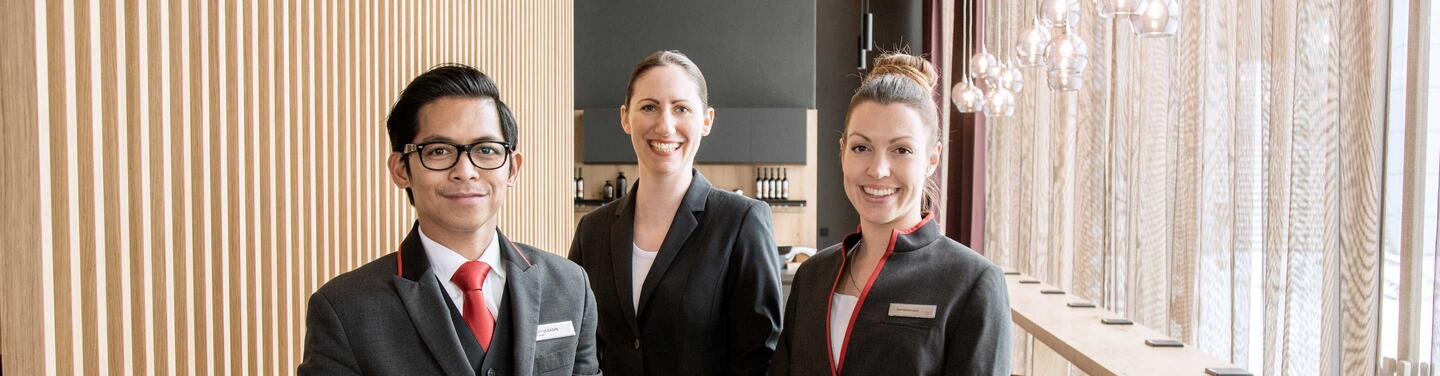 Mitarbeiter im Hilton Hotel Drei Personen in Serviceuniform stehen nebeneinander, lächeln in die Kamera und befinden sich in einem hellen, modernen Gastronomieumfeld.