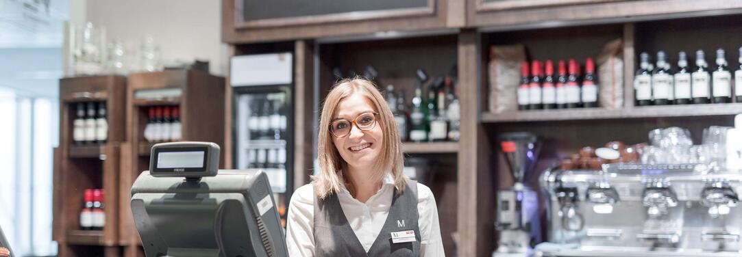 Allresto Trainee Junger Mensch im Servicebereich steht hinter einer Kasse vor Regalen mit Getränken und blickt freundlich in die Kamera.