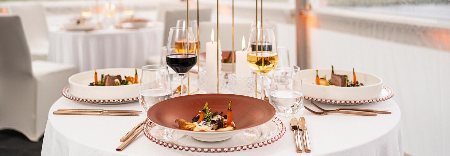 Zeltevent Catering Dinner Fein gedeckter Tisch mit edlen Weingläsern, goldenem Besteck und einem kunstvoll angerichteten Gericht auf einem roten Teller. Die elegante Dekoration vermittelt hochwertige Fine-Dining-Atmosphäre und exklusive Gastronomie am Flughafen München.