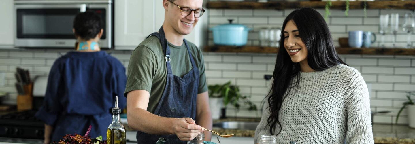 Kochkurse Zwei junge Erwachsene arbeiten gemeinsam lächelnd in einer modernen Küche, während im Hintergrund weitere Personen kochen. Die Szene strahlt Geselligkeit, Freude am Kochen und kreative Teamarbeit aus.