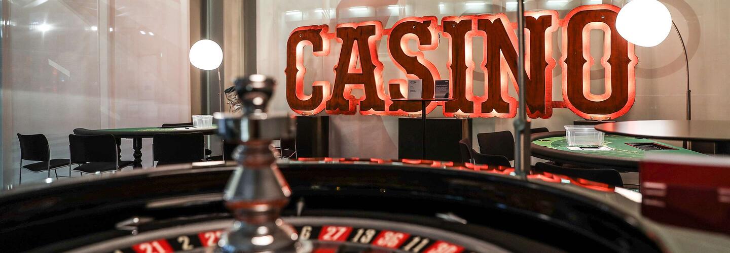 Themenevents Ein beleuchtetes ‚Casino‘-Schild thront über einem Roulette-Tisch im Vordergrund. Die warmen Lichter, glänzenden Oberflächen und das klassische Spielflair erzeugen eine aufregende, glamouröse Casino-Atmosphäre.