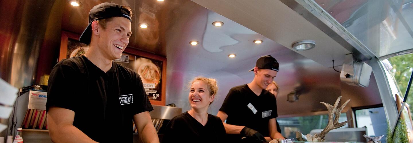 Streetfood Subralott Mehrere Mitarbeitende in einem modernen Foodtruck am Flughafen München arbeiten lachend zusammen. Die lockere, freundliche Stimmung und die offene Küche spiegeln authentisches Street-Food-Feeling und Teamspirit wider.