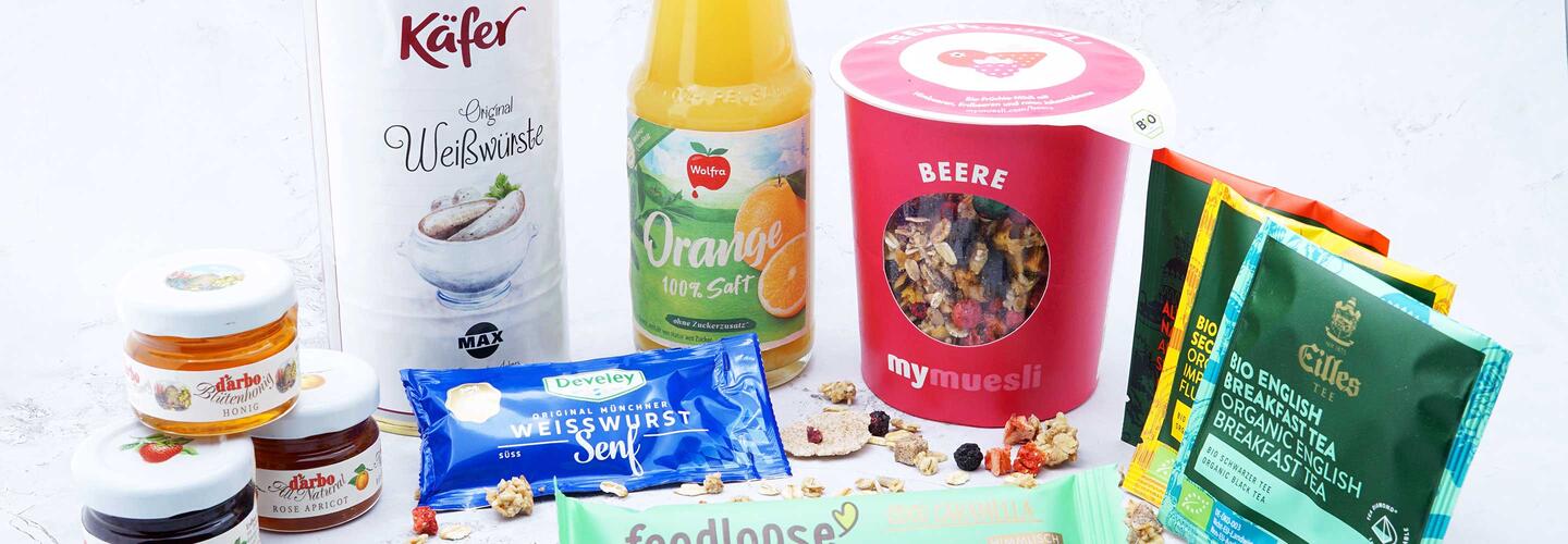 Frühstücksbox Vielfältiges Sortiment aus Snacks, Getränken, Süßwaren und Convenience-Produkten, darunter Marken wie Käfer und regionale Angebote. Die helle Präsentation vermittelt Frische und Auswahlvielfalt für Reisende am Flughafen München.