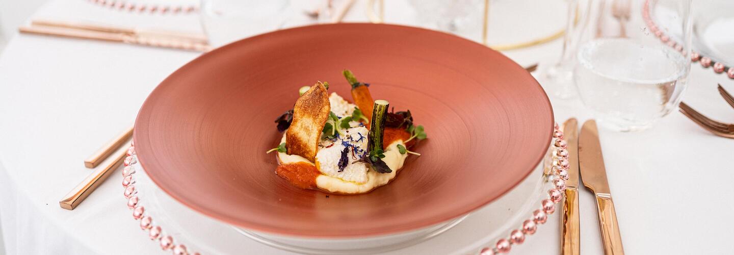 Menü Kunstvoll angerichtetes Gourmetgericht auf einem matten, rötlichen Teller: Zarte Komponenten aus Gemüse, Kräutern und einer knusprigen Komponente liegen dekorativ arrangiert. Die helle Tischdekoration mit Glas und Besteck schafft eine elegante, ruhige Fine-Dining-Atmosphäre.