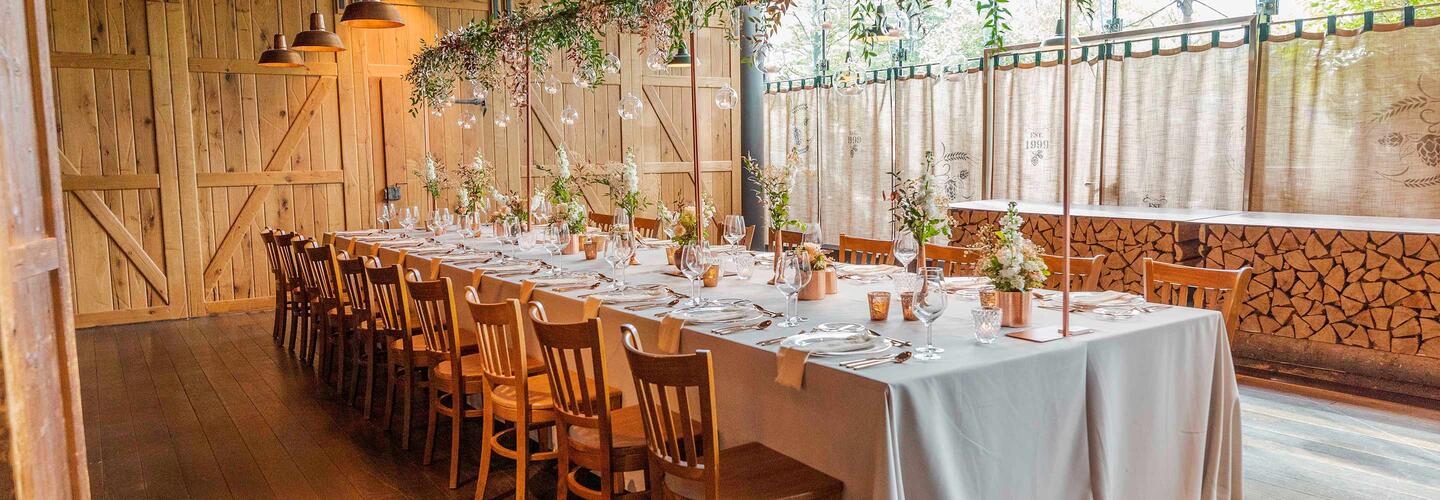 Airbräu Tenne Lange, festlich gedeckte Tafel in einer Scheunen- oder Almhütten-Location mit Holzwänden und gestapeltem Kaminholz. Dezente florale Dekoration und warmes Licht erzeugen eine gemütliche, natürliche und dennoch stilvolle Feieratmosphäre.