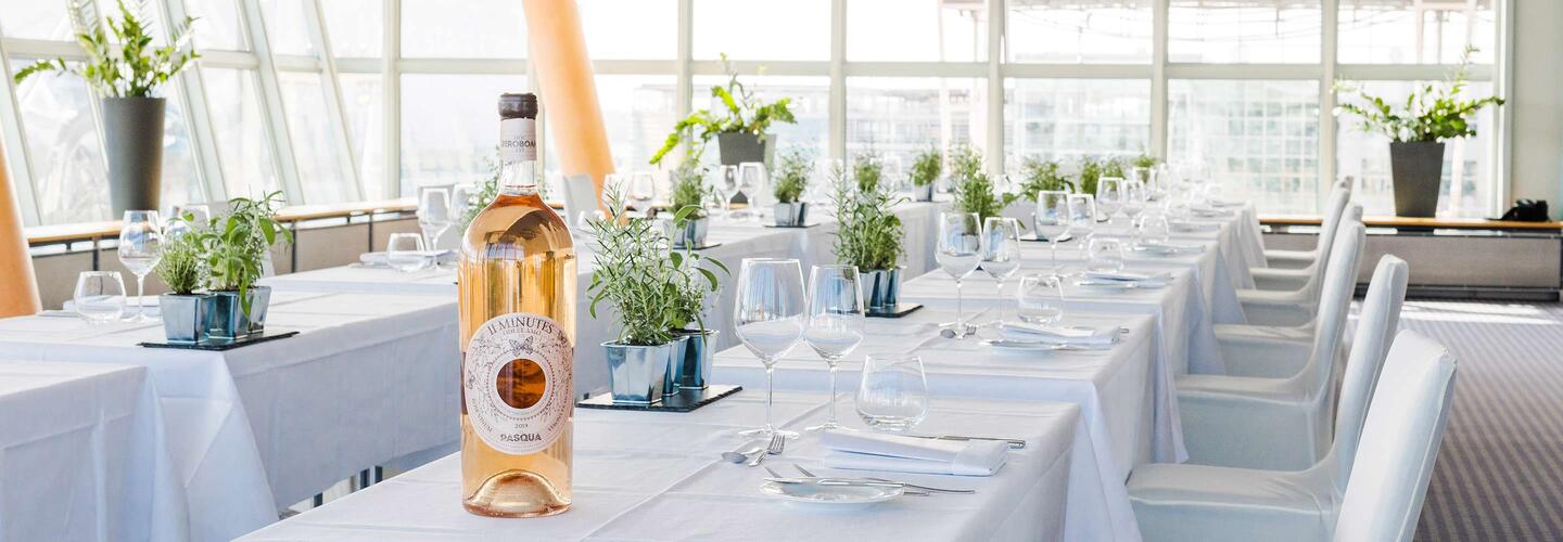 Location municon Lange Tafel mit weißen Stühlen und weißer Tischwäsche, dekoriert mit zarten Blumenarrangements in Glasvasen im municon. Eine Flasche Roséwein steht zentral auf dem Tisch. Große Fenster lassen viel Tageslicht herein und schaffen eine ruhige, festliche Atmosphäre.