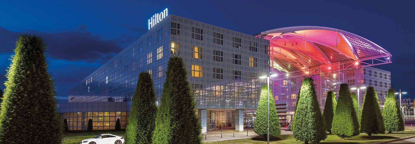 Hilton Hotel Das Hilton Hotel am Flughafen München leuchtet in der Dämmerung mit moderner Architektur, klaren Linien und einer spektakulären, pink-violett beleuchteten Überdachung. Gepflegte, konisch geschnittene Bäume rahmen den Eingangsbereich, die Szene wirkt hochwertig, urban und elegant.