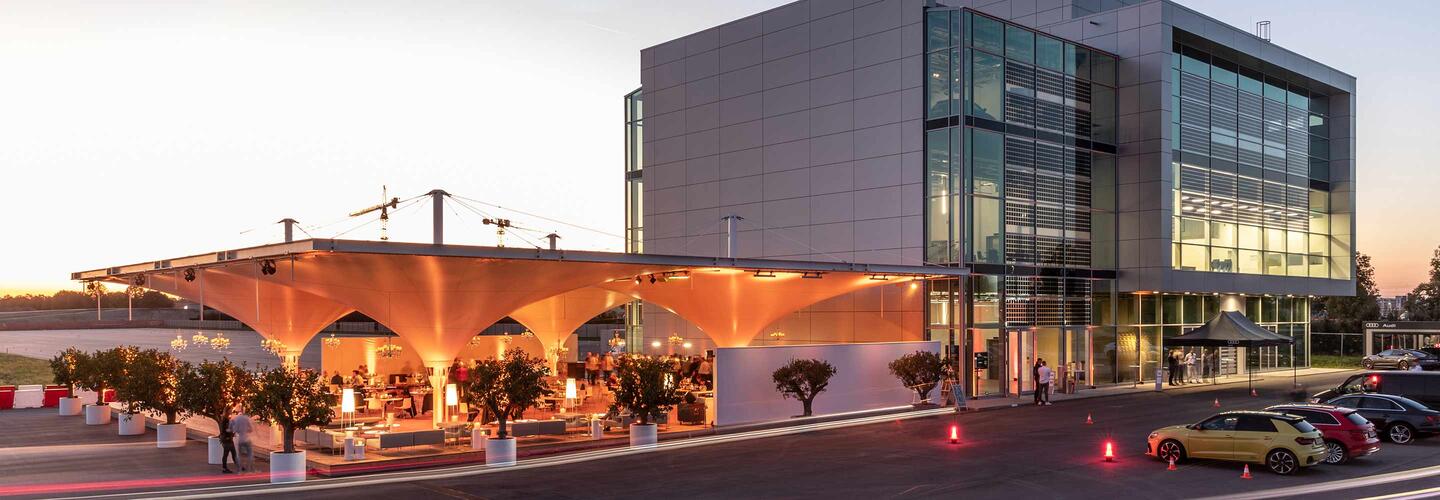 Audi Location Ein glasfrontiertes, modernes Gebäude am Flughafen München bei Sonnenuntergang, davor eine große überdachte Außenterrasse mit warmem Licht und Gästen. Die Stimmung ist einladend, lebhaft und zugleich stilvoll.