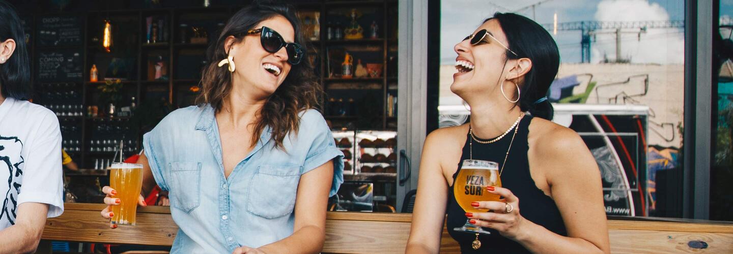 Privat Zwei junge Frauen sitzen lachend an einer Bar und halten bunte Getränke in der Hand. Sonnenbrillen, sommerliche Kleidung und das helle, urbane Ambiente vermitteln eine unbeschwerte, fröhliche Stimmung.