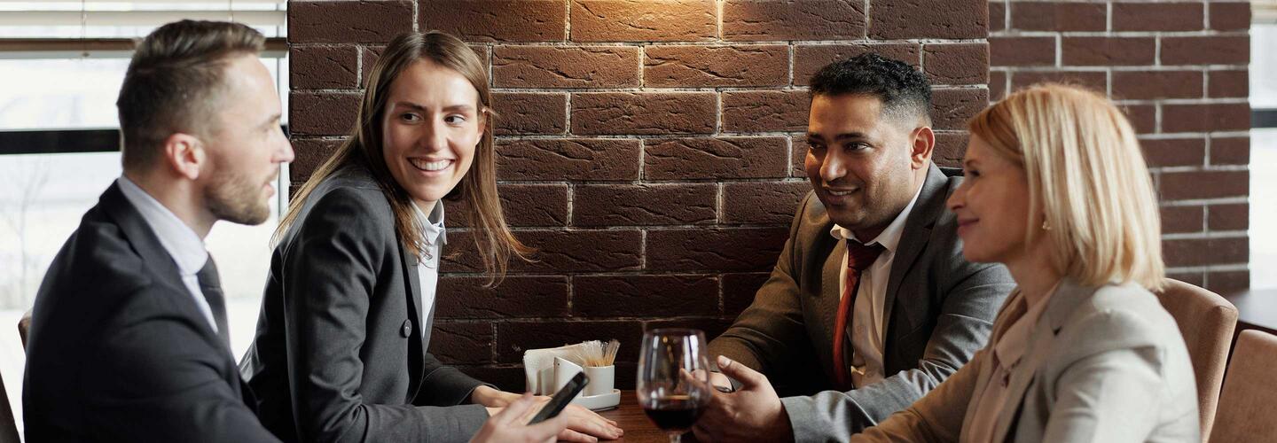 Business Vier Geschäftsleute sitzen an einem Holztisch, trinken Kaffee und führen ein angeregtes Gespräch. Die warme Beleuchtung und das moderne Café-Ambiente schaffen eine professionelle, aber zugleich entspannte Atmosphäre.