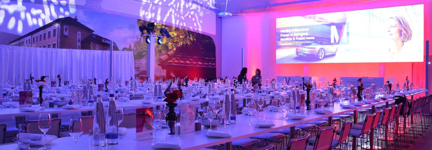 Firmenfeier Großer Veranstaltungssaal mit festlich gedeckten Rundtischen, weißer Tischdekoration und elegantem Ambiente. Farbige Beleuchtung in Pink und Violett taucht den Raum in eine glamouröse, moderne Atmosphäre. Im Hintergrund läuft eine große Präsentation auf einer Leinwand.