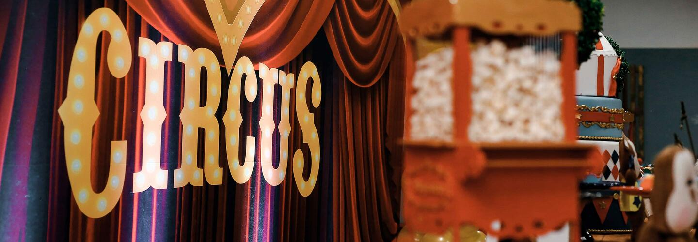 Zirkus Nostalgisches Circus-Schild in großen, beleuchteten Buchstaben hängt vor einem schweren roten Vorhang. Daneben steht ein Popcornstand. Die Szene wirkt verspielt, bunt und erinnert an klassische Zirkus- oder Jahrmarktsstimmung