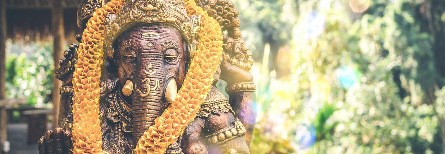 Oriental Detailaufnahme einer goldfarbenen Statue mit kunstvoll verziertem Kopf und Blumenkranz. Das weiche Sonnenlicht und das grüne, unscharfe Hintergrundlaub erzeugen eine ruhige, spirituelle und feierliche Stimmung.