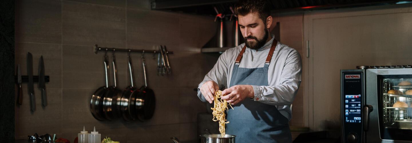Chefs Table Ein Koch in Schürze und Hemd hebt frische Pasta mit einer Zange aus einem Topf. Die professionell ausgestattete Küche im Hintergrund vermittelt handwerkliches Können, Konzentration und kulinarische Leidenschaft.
