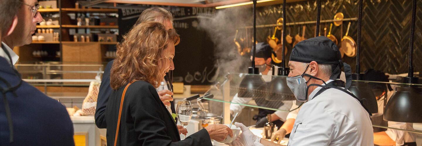 Walking Dinner Restaurantgäste stehen an einer offenen Kochstation und sehen einem Koch zu, der über dampfenden Töpfen arbeitet. Die Szene wirkt warm, einladend und zeigt authentische, handgemachte Küche in lebendiger Atmosphäre.