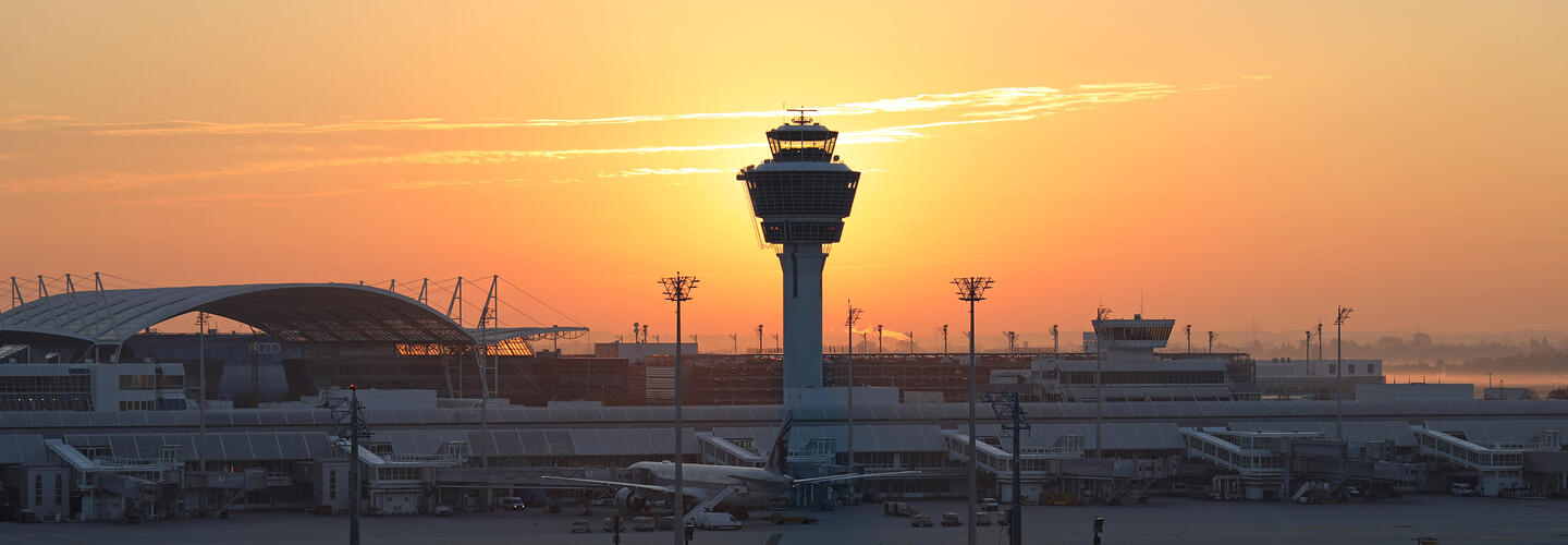 Panoramaansicht des Flughafens München Der Tower des Flughafens München ragt vor einem intensiven orangefarbenen Abendhimmel auf. Silhouetten von Terminaldächern und Beleuchtungsmasten zeichnen sich gegen das warme Licht ab. Die Stimmung ist ruhig, atmosphärisch und leicht melancholisch.