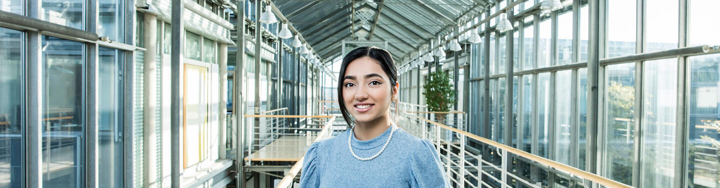 Ausbildung Eine Auszubildende steht lächelnd in einem lichtdurchfluteten Glasgang am Flughafen München. Das Motiv symbolisiert moderne Ausbildungsberufe, persönliche Entwicklung und vielfältige Karrierechancen in einem internationalen Arbeitsumfeld.