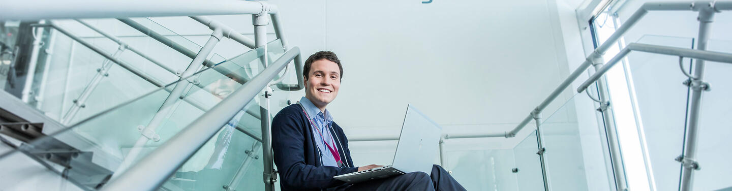 Duales Studium Junger Studierender sitzt mit Laptop auf einer Glastreppe im modernen Flughafengebäude und lächelt in die Kamera.