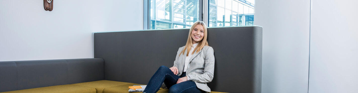 Schülerpraktikum Eine junge Praktikantin sitzt entspannt auf einer modernen Sitzbank im Flughafengebäude und lächelt in die Kamera. Das Bild symbolisiert das Schülerpraktikum am Flughafen München, bei dem Jugendliche wertvolle Einblicke in verschiedene Berufsfelder erhalten und erste praktische Erfahrungen sammeln können.