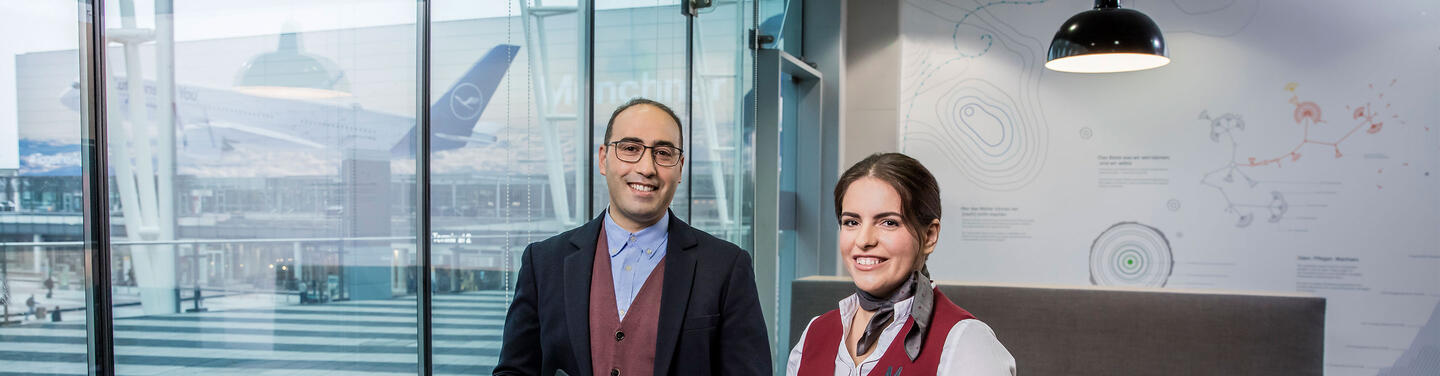 Berufseinsteiger, Berufserfahrene & Quereinsteiger Zwei Berufseinsteiger stehen lächelnd in einem modernen Empfangsbereich mit Blick auf das Terminal und ein Flugzeug des Flughafens München.