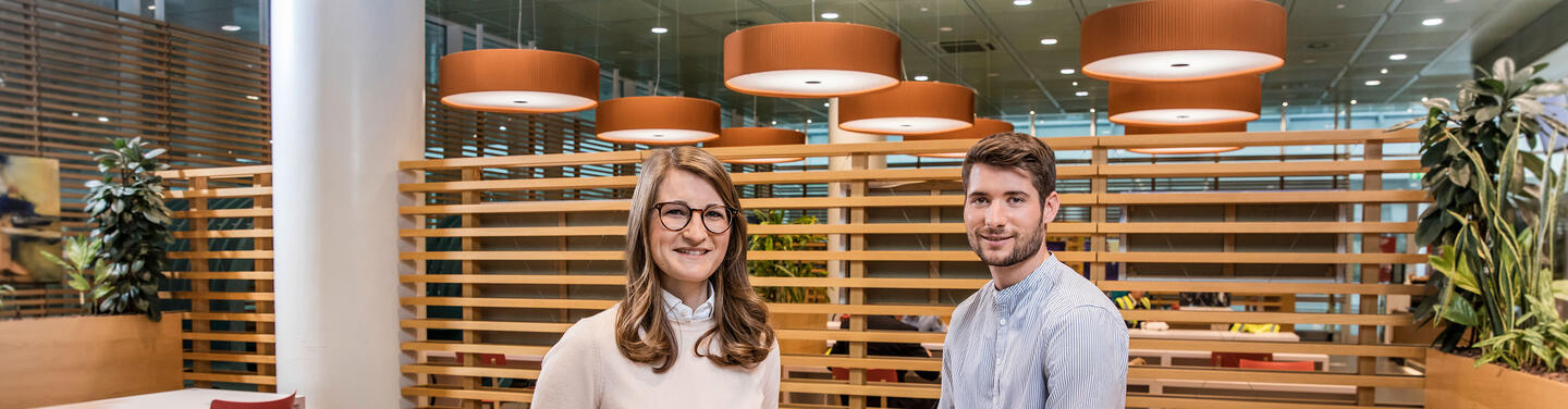 Trainee Zwei Hochschulabsolvierende stehen lächelnd in einem modernen Arbeitsumfeld mit Holztrennwänden und orangefarbenen Deckenleuchten am Flughafen München.