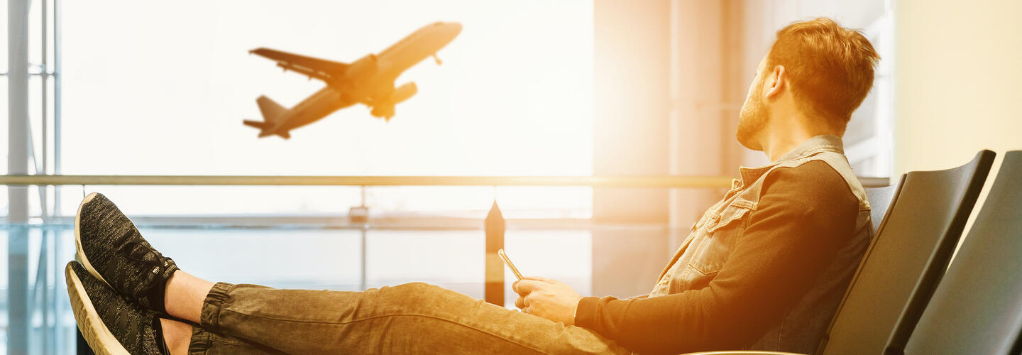 Gate Catering Ein Mann sitzt entspannt an einem Gate-Fenster, blickt auf das Rollfeld und beobachtet ein startendes Flugzeug, dessen Silhouette sich gegen das helle Sonnenlicht abzeichnet. Die Szene wirkt ruhig, reflektiert und weckt Fernweh.