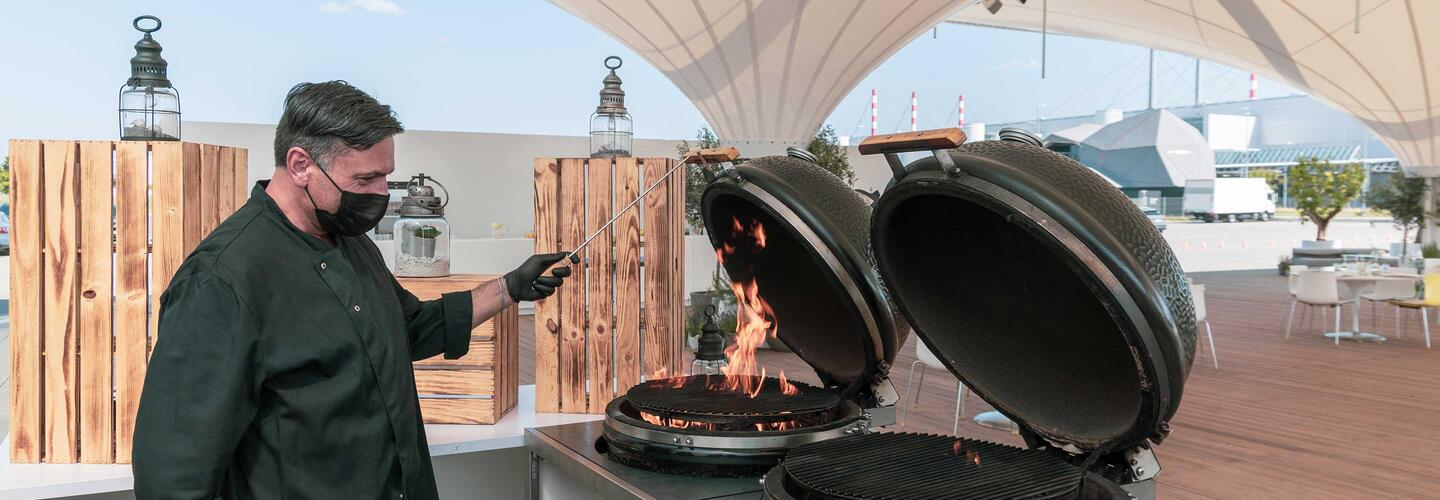 BBQ Ein Koch steht auf einer Terrasse vor einem großen Keramikgrill, dessen Deckel geöffnet ist und aus dem Flammen lodern. Im Hintergrund spannen sich helle Sonnensegel über den Außenbereich. Die Szene wirkt lebhaft, rustikal und zeigt die Vorfreude auf frisch Gegrilltes.