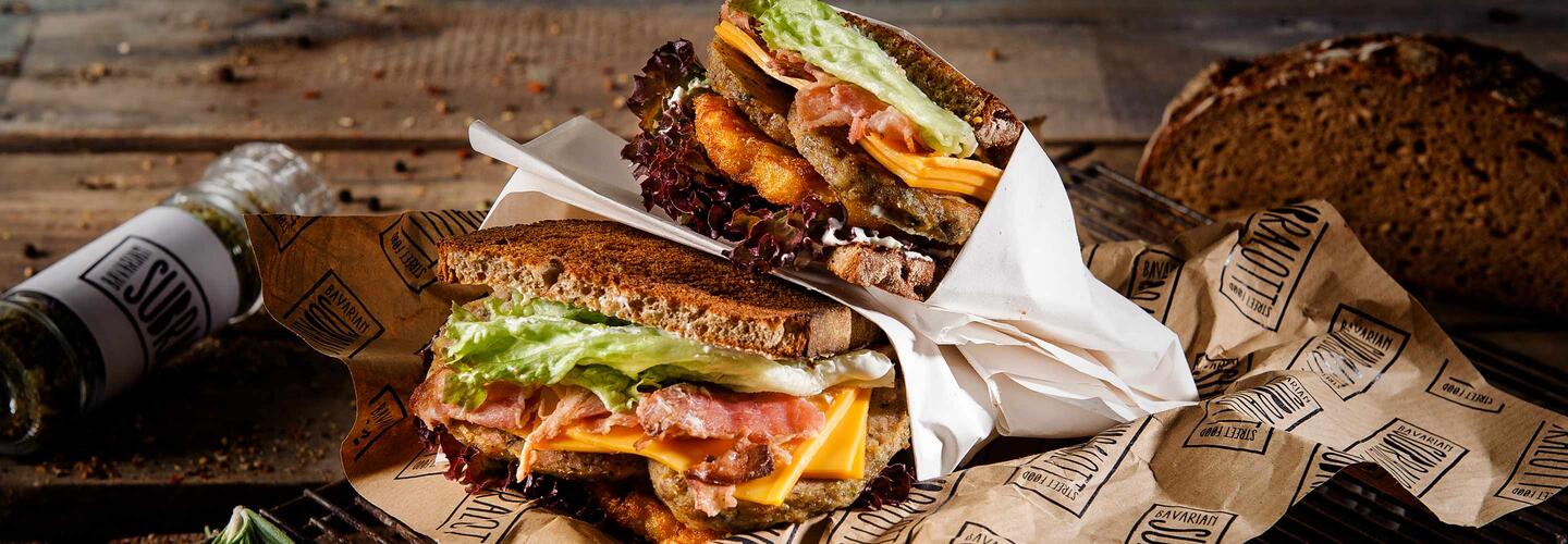 Subralott Sandwich Zwei belegte Sandwiches mit knusprigem Brot, Fleisch, Käse und Gemüse liegen auf Papier serviert, umgeben von einer Flasche und rustikalem Tischzubehör. Die Stimmung ist herzhaft, warm und appetitanregend – ein echter Street-Food-Charakter.