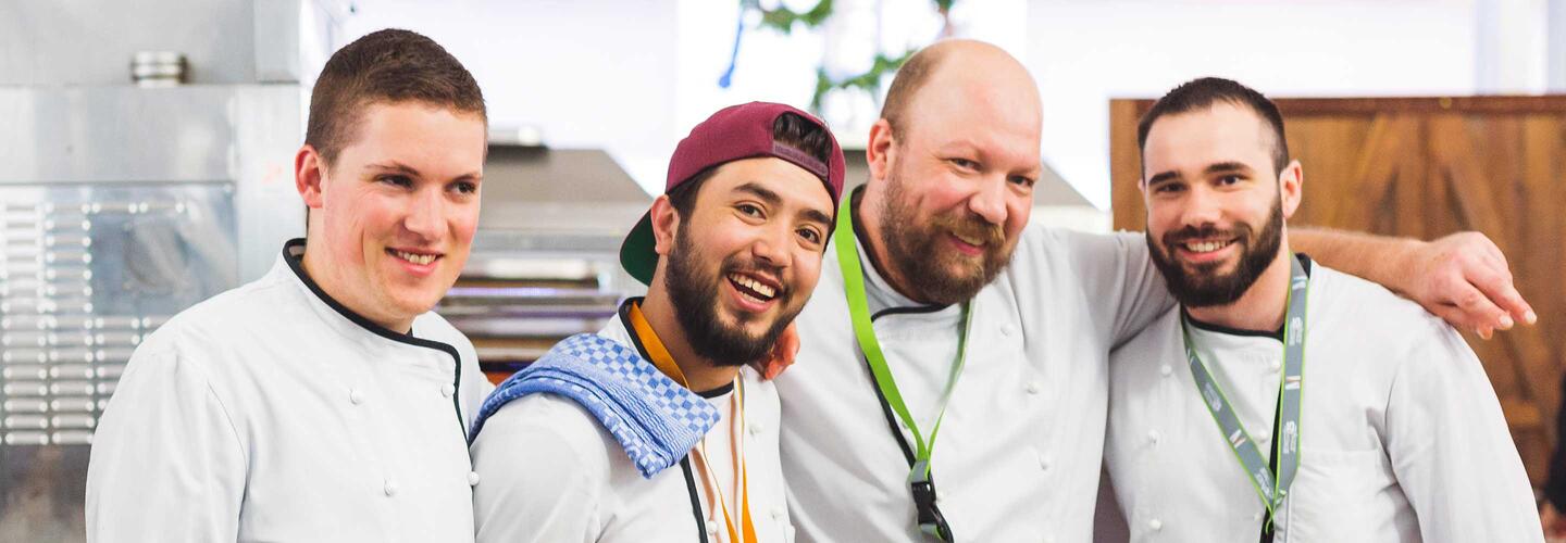 Über uns Vier junge Köche stehen eng nebeneinander in einer professionellen Küche und lächeln in die Kamera. Ihre fröhliche, offene Körperhaltung vermittelt Teamgeist, Freude am Beruf und eine freundliche Arbeitsatmosphäre.