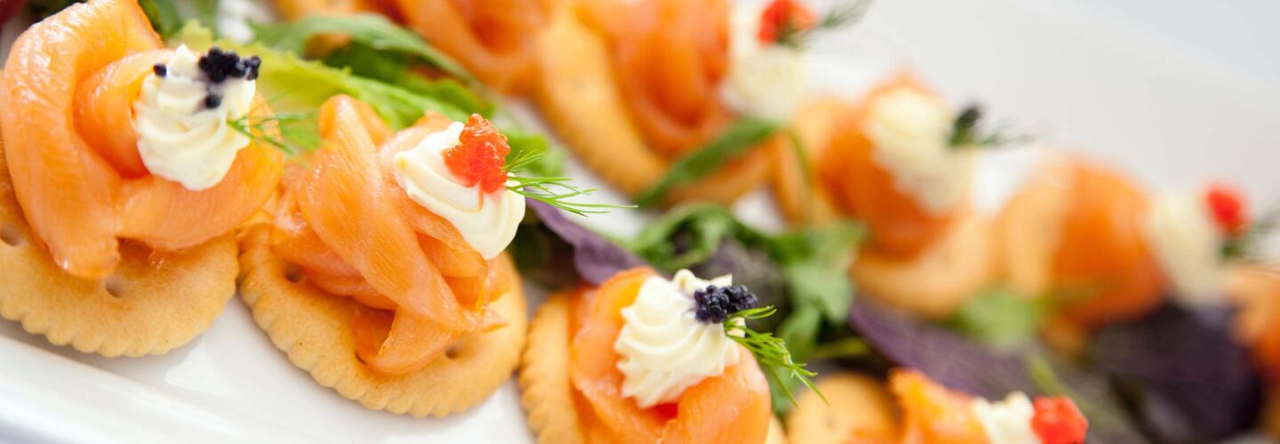 Fingerfood Fein dekorierte Lachs-Häppchen mit Creme, Kräutern und kleinen Toppings liegen kunstvoll auf einer weißen Platte. Die zarte Farbgebung und detailreiche Anrichtung erzeugen einen hochwertigen, appetitlichen Eindruck.