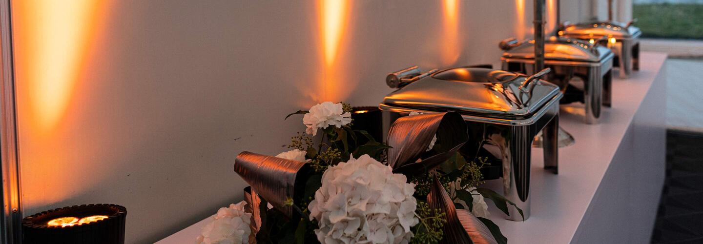 Catering-Buffet Stilvoll angerichtetes Catering-Buffet mit silbernen Warmhaltebehältern und weißer Blumendekoration, beleuchtet von warmen, goldenen Wandstrahlern. Die Atmosphäre ist festlich, hochwertig und einladend.
