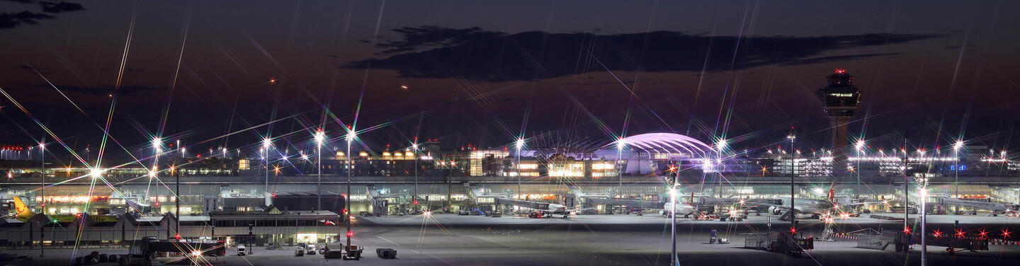 Keyvisual Terminal Night Night-time view of a brightly illuminated airport terminal with aircraft parked at the gates, numerous light reflections on the apron, and the control tower visible in the background; the scene appears lively, clear, and atmospheric.