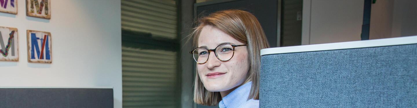 Duales Studium Lächelnde Mitarbeiterin mit Brille sitzt an einem Büroarbeitsplatz mit grauer Trennwand in heller, moderner Umgebung.