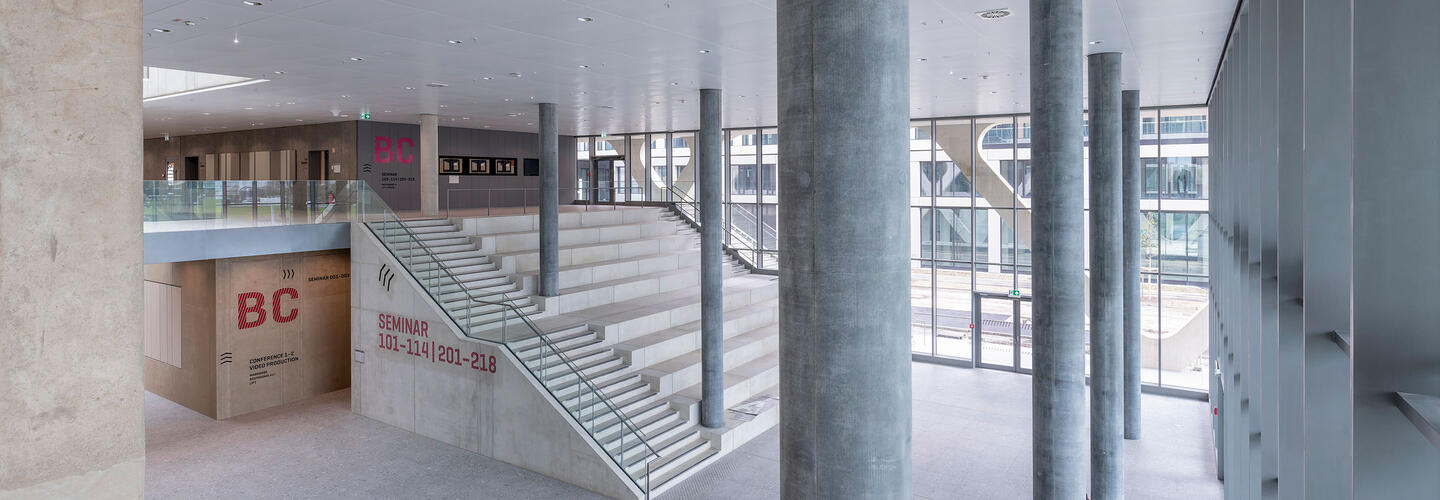 AirportAcademy - LAB 52 Bright, open entrance hall with wide staircases, large windows, and clean lines made of concrete and glass. The calm atmosphere conveys order, structure, and modern design.