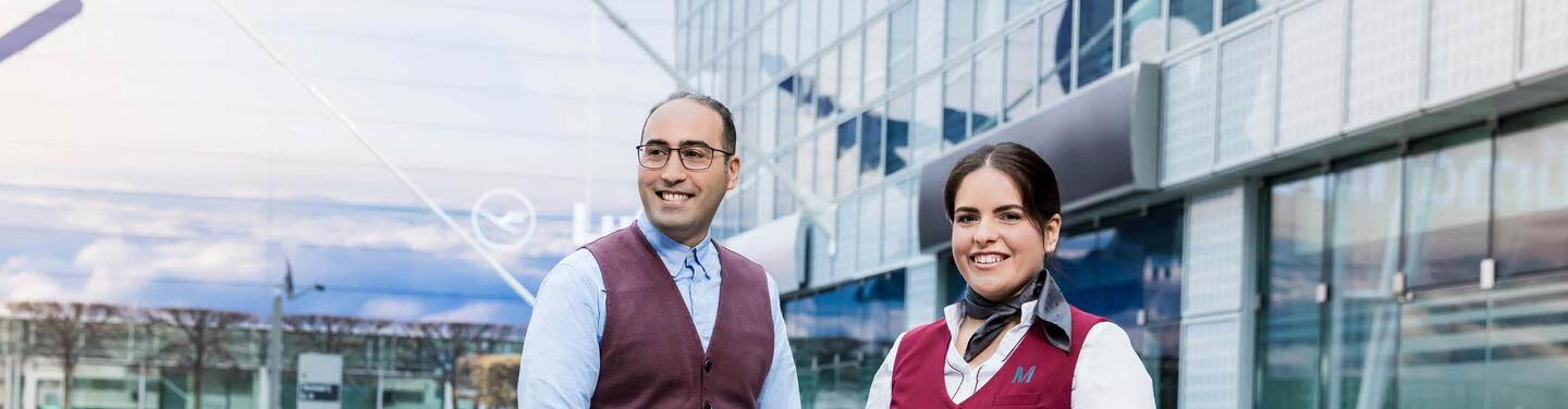 Flughafen live: Karriertag Zwei Mitarbeitende des Flughafens München stehen lächelnd vor einem modernen Glasgebäude.